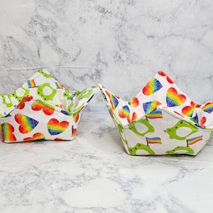 May include: Two fabric bowl cozies featuring a white background with a repeating pattern of green frogs holding rainbow flags and colorful rainbow-textured hearts. Each square-shaped cozy has four pointed corners and a quilted texture.