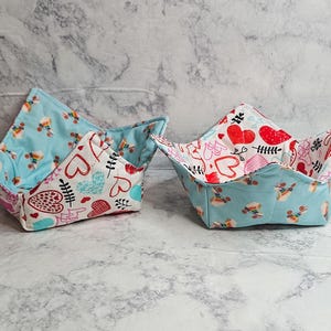 May include: Two fabric bowl cozies with a square, four-cornered shape. One cozy features a light blue interior with a roller skate pattern and a white exterior with red and blue heart motifs. The second cozy reverses these fabric patterns.