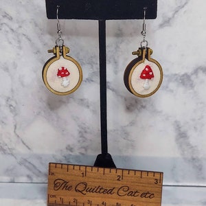 May include: Embroidered mushroom earrings. Each earring features a small, round wooden hoop with a cream-colored fabric background. A red mushroom with white spots is embroidered on the fabric. The earrings hang from silver-colored hooks. A wooden ruler is in the foreground.