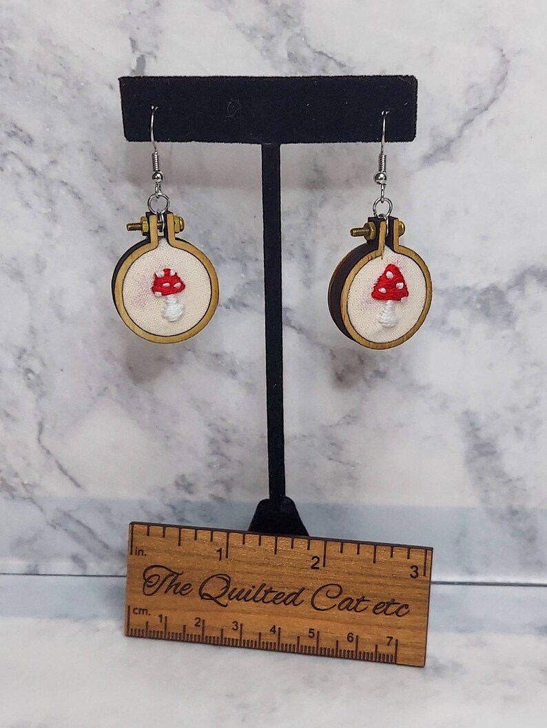 May include: Embroidered mushroom earrings. Each earring features a small, round wooden hoop with a cream-colored fabric background. A red mushroom with white spots is embroidered on the fabric. The earrings hang from silver-colored hooks. A wooden ruler is in the foreground.