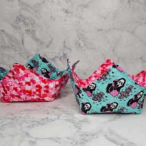 May include: Two fabric bowls with a unique design. One bowl features a pink and red heart pattern, while the other has a teal background with a black and white ghost graphic. The bowls are folded into a square shape.