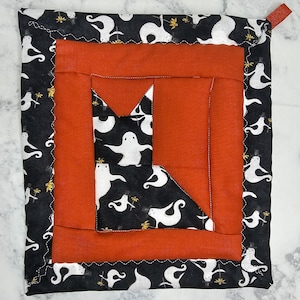 May include: A square quilted potholder featuring a central cat silhouette pieced from black fabric with a white ghost and yellow flower pattern, framed by a solid orange border and an outer black border with the same ghost and flower print.