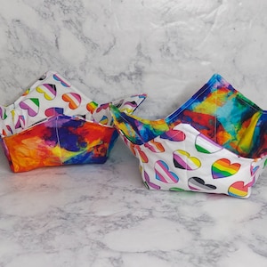 May include: Two fabric bowls with a white background and colourful hearts. The hearts are printed with different pride flag designs. The bowls are folded and ready to use.
