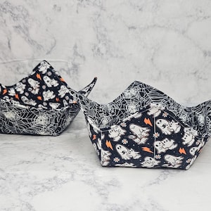 May include: Two fabric bowl cozies featuring a reversible design. One side displays a black background with white ghosts, orange lightning bolts, and small flowers. The other side features a black and white spider web pattern throughout the fabric.