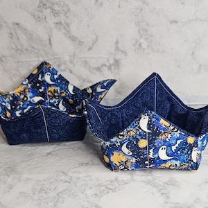 May include: Two Halloween bowl cozies. The exterior is a dark blue fabric with a swirl pattern, while the interior features a spooky print with ghosts, bats, and moons on a blue background. The cozies are designed to hold bowls.