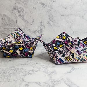 May include: Two fabric bowl cozies featuring a reversible design. One side displays a dark background with white mice, yellow stars, and pink flowers. The other side features a light background with colorful books, rainbow stripes, and green pine trees.