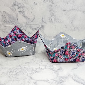 May include: Two fabric bowl cozies with four-cornered square designs. One cozy features a gray exterior with white daisy prints and a dark floral interior. The second cozy has a dark floral exterior and a gray interior with white daisy prints.