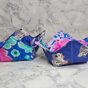 May include: Two fabric bowl cozies featuring a blue base with a floral pattern and a print of a white animal with the text "YOU SHOULD SMILE MORE". The bowl holders have a square, quilted design with four pointed corners and a vibrant, colorful aesthetic.