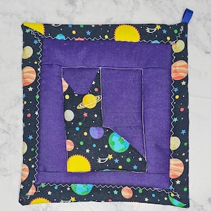 May include: Square quilted potholder with a purple center and a black border featuring planets, stars, and yellow suns. A section of the potholder has a black background with planets and stars. A blue loop is attached for hanging.