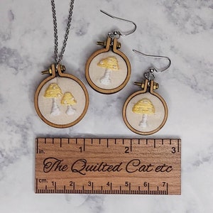 May include: A handmade jewelry set featuring embroidered mushroom designs. The set includes a necklace and a pair of earrings, each set in a small wooden hoop. The necklace has a silver chain. The ruler shows measurements in inches.