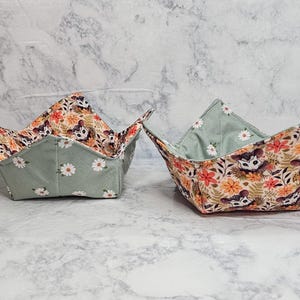 May include: Two fabric bowl cozies featuring a reversible design. One side displays a pattern of opossums, orange and yellow flowers, and green ferns. The other side is a solid sage green fabric decorated with a repeating pattern of white daisies.