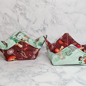 May include: Two fabric bowl cosies, one brown with mushrooms and one green with a woodland scene. Both cosies are folded into a bowl shape.