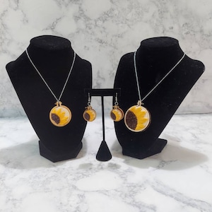 May include: A set of sunflower earrings and a necklace. The earrings and necklace feature a yellow sunflower with brown center on a round wooden base.