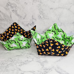 May include: Two Halloween-themed fabric bowls. One bowl features a black exterior with candy corn, and the other has a green exterior with ghosts and bats. Both bowls have a green interior with ghosts and bats.