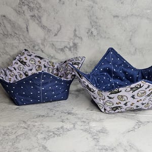 May include: Two fabric bowl cozies. The exterior is navy blue with white polka dots, and the interior features a light purple pattern with bats, cats, and moons. The cozies are designed to hold bowls.