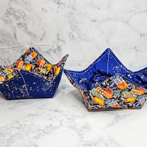 May include: Two fabric bowl cozies featuring a dark blue exterior with gold speckles and an interior patterned with orange and black Dungeon Crawler text and graphics. The bowl holders have a square base and four pointed corners.