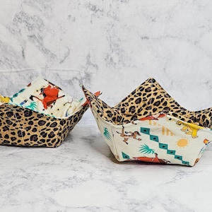 May include: Two fabric bowl cozies. One features a leopard print exterior and a patterned interior with cartoon characters. The other has a leopard print exterior and a patterned interior with cartoon characters. The cozies are designed to hold bowls.