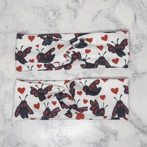 May include: Two white fabric headbands with a knot in the center. The headbands have a repeating pattern of red hearts and black moths with red eyes.