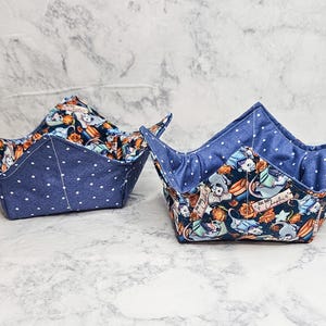 May include: Two fabric bowl cozies featuring a dark blue exterior with white polka dots and an interior pattern of illustrated opossums reading books, orange flowers, and the text "Big Book Chapter 4" printed on a rectangular label.