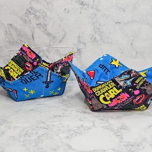 May include: Two fabric bowl cozies featuring a vibrant print with the text Dungeon Crawler Carl, pixelated swords, and neon graphic elements. One cozy has a blue exterior and black interior, while the other has a black exterior and blue interior.