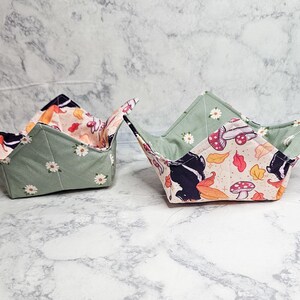May include: Two fabric bowl cozies. The exterior is sage green with white daisy patterns. The interior features a whimsical print with mushrooms, leaves, and a black and white skunk silhouette. These cozies are designed to hold bowls.