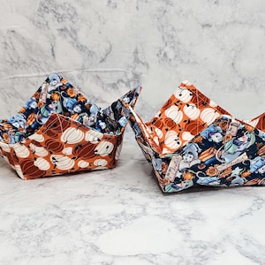 May include: Two fabric bowl cozies featuring a reversible design. One side displays a pattern of white and orange pumpkins on an orange background, while the other side features a dark blue print of small animals reading books and orange flowers.