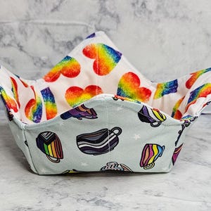 May include: A fabric bowl cozy featuring a light blue exterior with a pattern of various colorful mugs and small white stars. The interior lining is white fabric decorated with a repeating pattern of vibrant rainbow-colored hearts.