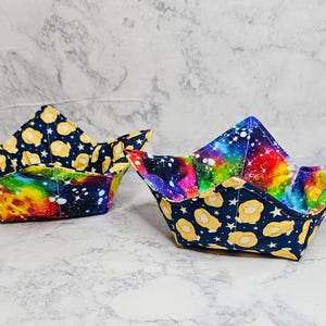 May include: Two fabric bowl cozies. One has a navy blue exterior with yellow egg and star patterns, and a rainbow interior. The other has a navy blue exterior with yellow egg and star patterns, and a galaxy-themed interior.