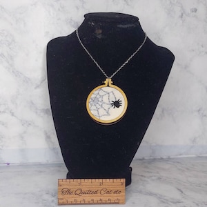 May include: A black and white spider web embroidery on a round wooden pendant, hanging from a silver chain necklace. The pendant is 2 inches in diameter.