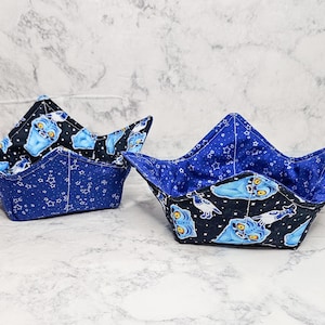 May include: Two fabric bowl cozies. The exterior is a dark blue with white star patterns, and the interior is black with light blue alien-like figures. The cozies are designed to hold bowls.