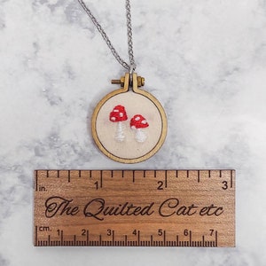 May include: A handmade necklace featuring a small, embroidered mushroom design. The mushrooms have red caps with white spots and are set in a wooden embroidery hoop. The necklace includes a silver chain. The wooden ruler below measures in inches and centimeters, with the text "The Quilted Cat etc."
