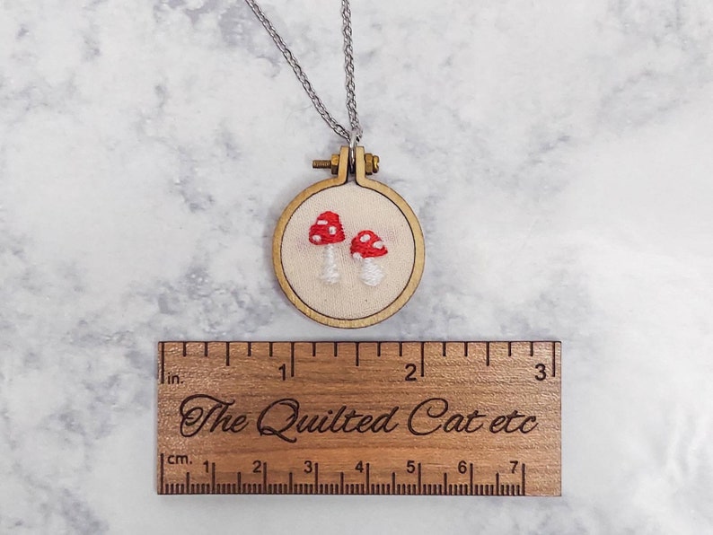 May include: A handmade necklace featuring a small, embroidered mushroom design. The mushrooms have red caps with white spots and are set in a wooden embroidery hoop. The necklace includes a silver chain. The wooden ruler below measures in inches and centimeters, with the text "The Quilted Cat etc."