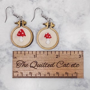 May include: A pair of earrings featuring miniature embroidery hoops with red and white mushroom designs on cream fabric. A wooden ruler is placed below the earrings, showing the hoops are approximately 1 inch in diameter. The ruler is engraved with the text The Quilted Cat etc.