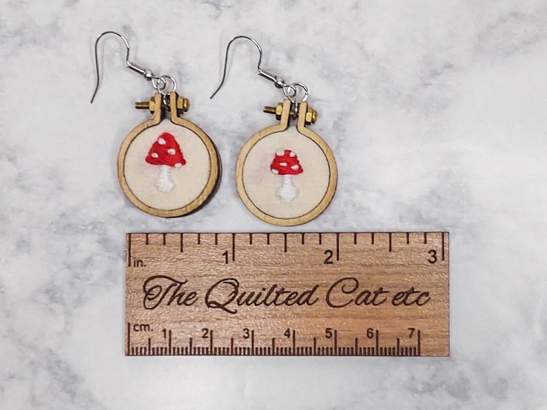 May include: A pair of earrings featuring miniature embroidery hoops with red and white mushroom designs on cream fabric. A wooden ruler is placed below the earrings, showing the hoops are approximately 1 inch in diameter. The ruler is engraved with the text The Quilted Cat etc.