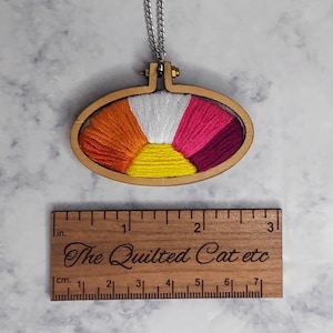 May include: A wooden oval embroidery hoop necklace with a rainbow flag design. The hoop is about 2 inches in diameter and has a gold chain.