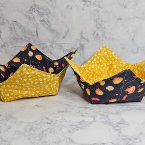 May include: Two handmade fabric bowl cozies. The exterior is yellow with a white floral pattern, and the interior is black with a cartoon character design. The bowl cozies are designed to hold hot bowls and protect hands.