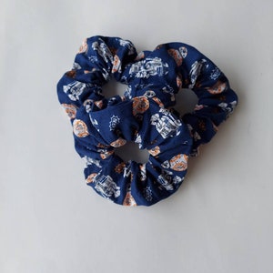 May include: Three navy blue scrunchies with a repeating pattern of white and orange robot illustrations. The fabric is gathered, creating a ruffled effect. The scrunchies are designed for hair styling.