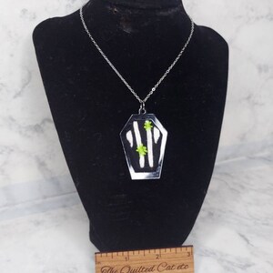 May include: A silver chain necklace with a black and white coffin pendant. The coffin has a green floral design and is about 2 inches long.