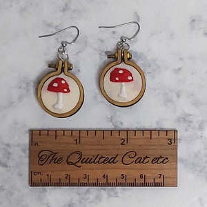 May include: A pair of earrings featuring miniature wooden embroidery hoops with white fabric and red mushroom embroidery. A wooden ruler is placed below, showing the earrings are approximately 1.25 inches in diameter.