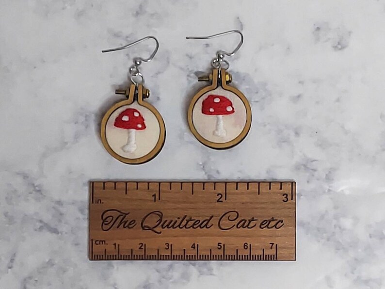 May include: A pair of earrings featuring miniature wooden embroidery hoops with white fabric and red mushroom embroidery. A wooden ruler is placed below, showing the earrings are approximately 1.25 inches in diameter.
