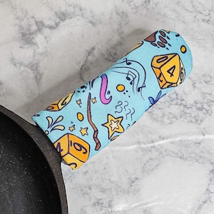 May include: A blue and yellow fabric pot holder with a dice and other whimsical designs. The pot holder is wrapped around a black cast iron skillet handle.