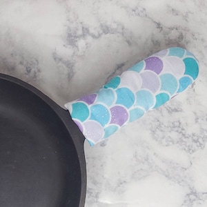 May include: A black cast iron skillet with a teal, purple, and white mermaid scale patterned oven mitt on the handle.