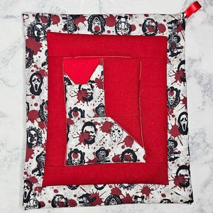 May include: Square red and white pot holder with a red loop. The pot holder has a red center and a border with horror movie character faces and red splatter designs.