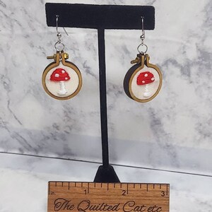 May include: A pair of handmade earrings featuring embroidered red and white mushrooms within small wooden embroidery hoops. The earrings hang from silver-colored hooks. The Quilted Cat etc is engraved on a wooden ruler.