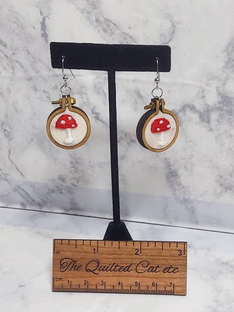 May include: A pair of handmade earrings featuring embroidered red and white mushrooms within small wooden embroidery hoops. The earrings hang from silver-colored hooks. The Quilted Cat etc is engraved on a wooden ruler.