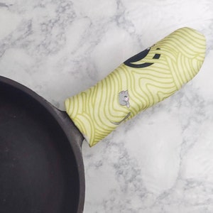 May include: A black cast iron skillet with a yellow and green patterned silicone handle. The handle has a cat design on it.