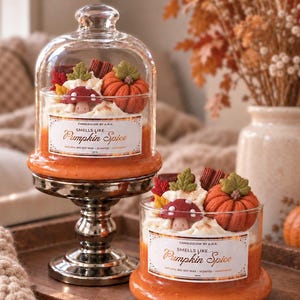 Könnte beinhalten: Zwei orangefarbene, nach Kürbisgewürz duftende Kerzen in Gläsern mit weißem Wachs-Topping in Form von Kürbissen, Pilzen und Blättern. Die Etiketten tragen die Aufschrift CANDLEGLOW BY A.N.A. SMELLS LIKE... Pumpkin Spice.