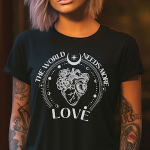 Könnte beinhalten: Schwarzes T-Shirt mit einem weißen Grafikdesign, das ein Herz mit Blumen und den Text "The World Needs More Love" zeigt.