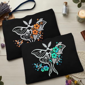Könnte beinhalten: Zwei schwarze Reißverschlusstaschen mit einem weißen Motten- und Blumendesign. Eine Tasche hat orangefarbene Blumen, die andere hat türkisblaue Blumen. Beide haben eine Handschlaufe. Die Taschen liegen auf einer hellen Holzoberfläche.