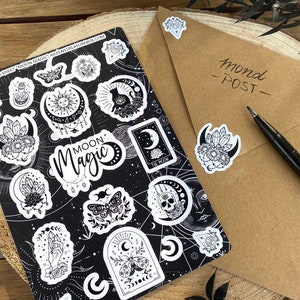 Puede incluir: Una hoja de pegatinas en blanco y negro con las palabras "Moon Magic" y varias ilustraciones con tem&aacute;tica lunar, de cristales y celestial. La hoja est&aacute; sobre una superficie de madera.